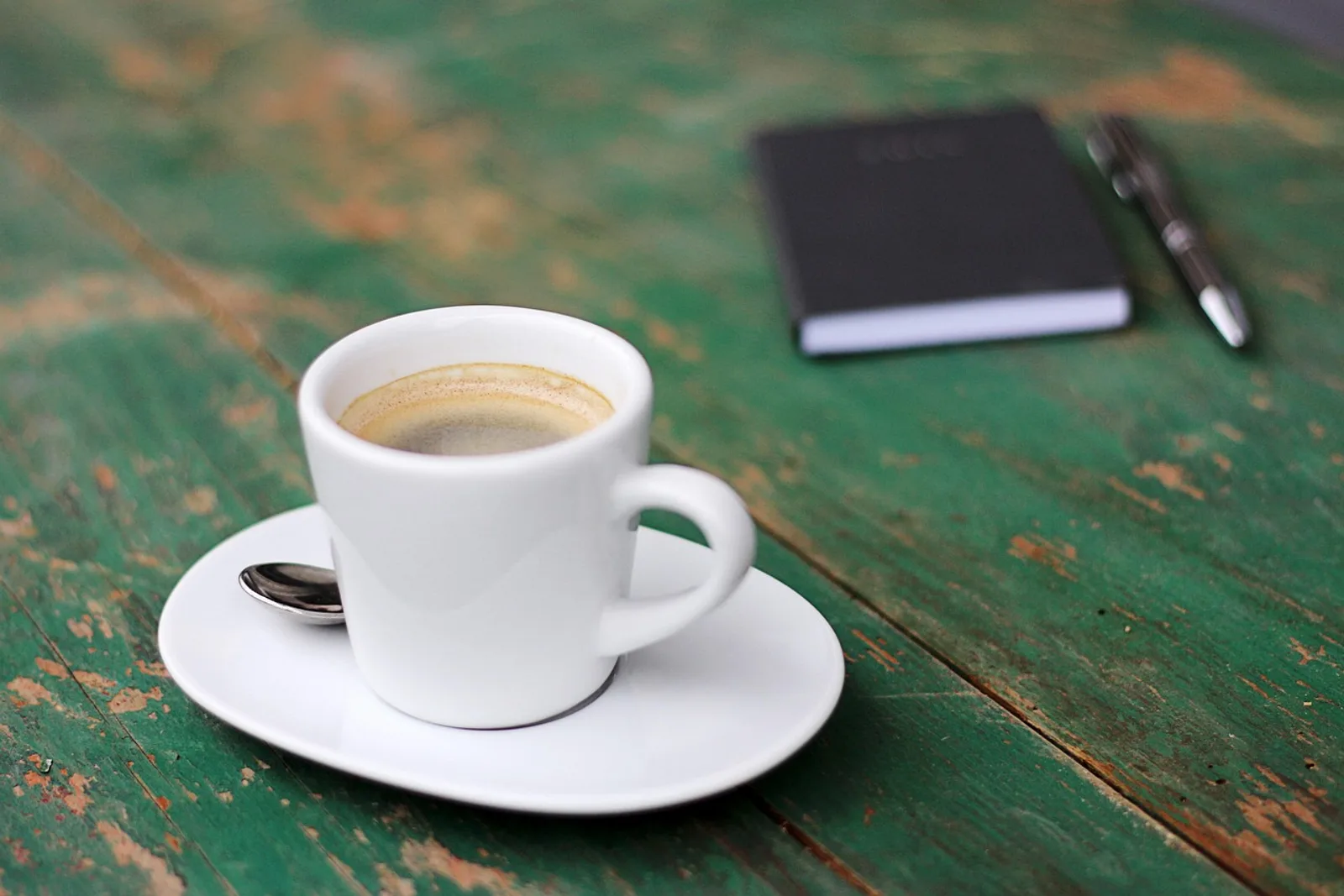 Espresso auf grünem Holztisch, dahinter ein geschlossenes Notizbuch mit Stift – Aufgaben priorisieren am Morgen