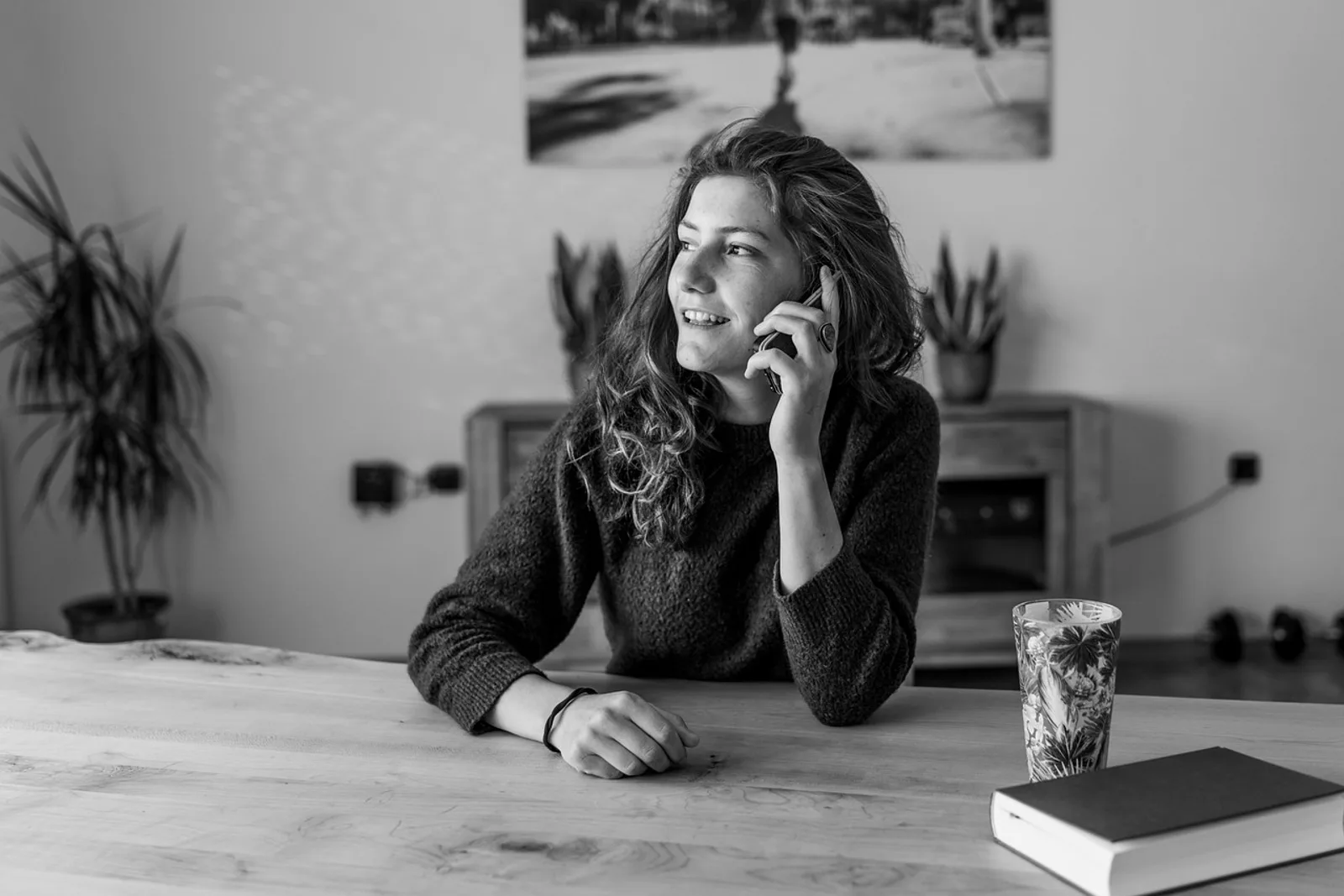Frau telefoniert lächelnd am Holztisch, daneben Glas und Buch – Austausch statt Vergleichen
